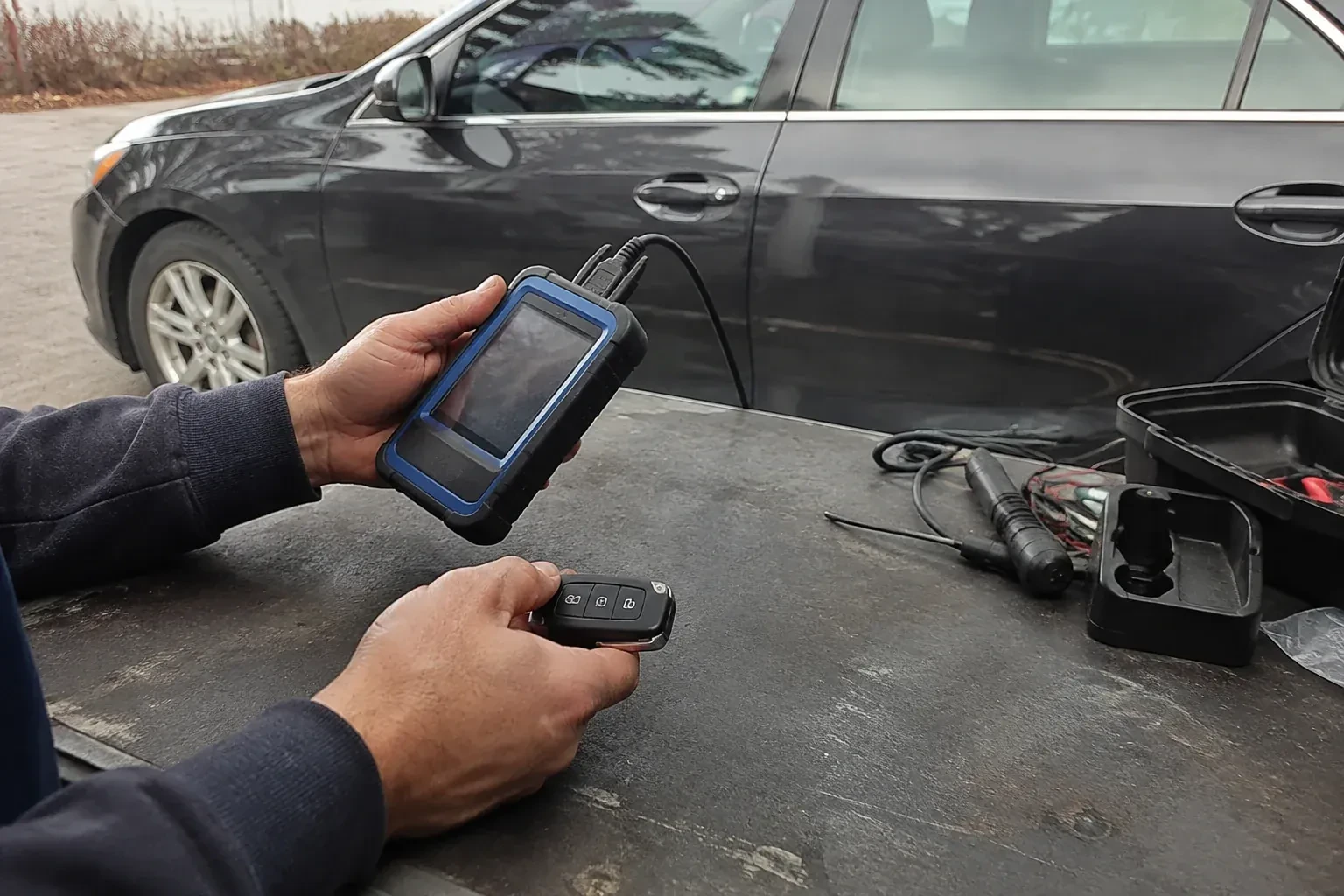 Hands holding a blue-edged electronic device and car-key fob at a worktable beside a parked car. — Transponder Key Programming, Brampton