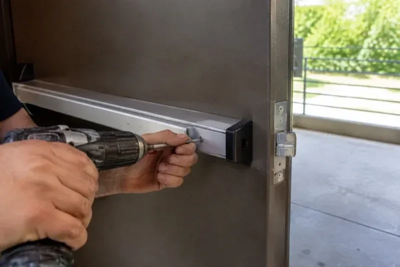 Hands using a power driver at the end of a horizontal push bar on an open metal door. — Panic & Exit Hardware, Brampton